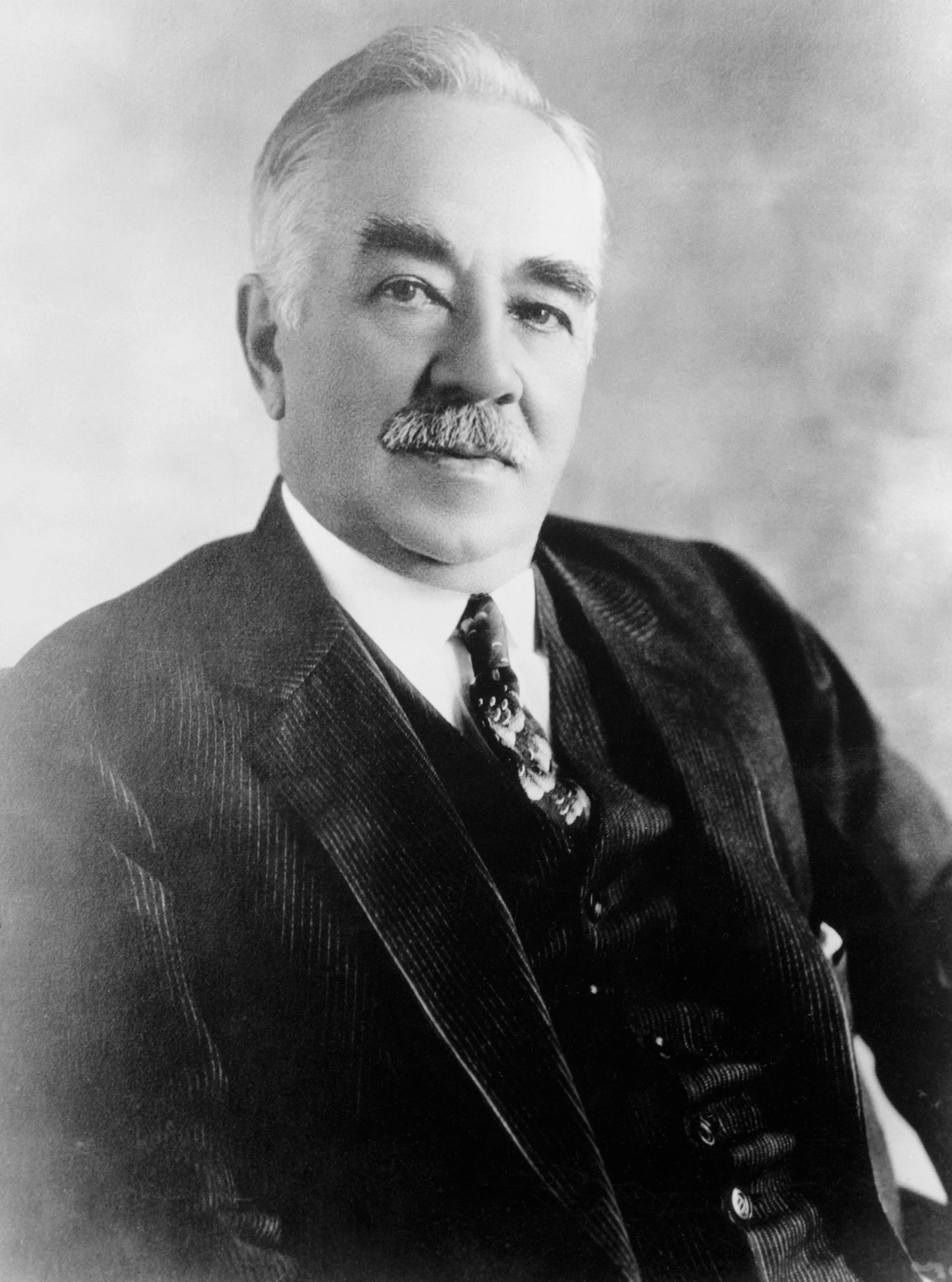 A man with white hair and a thick but trim moustache, in a three-piece suit, looks confidently at the camera in this black-and-white portrait.