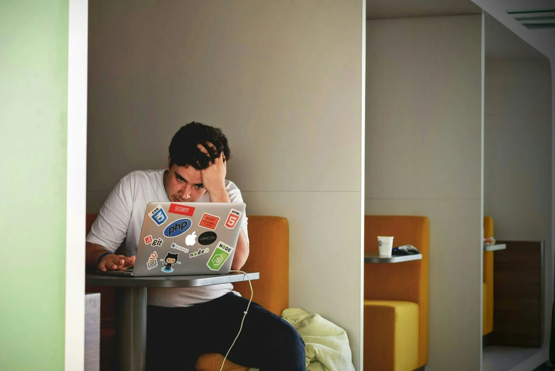 A young man in a white T-shirt stares into a stickered laptop at a coffee shop, head in hand.