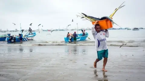 Getty Images A fisherman carries freshly caught tuna at La Poza beach in Manta, Ecuador's largest seaport. He is wearing a white football shirt and turquoise shorts, with bare feet as he walks across the wet sand.