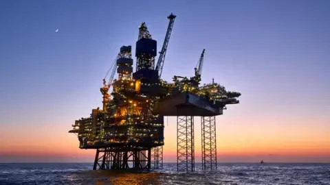 Getty Images An oil platform seen from below stands in the North Sea, the sun is setting and the moon can be seen. Lights are on across the platform which stands on stilts with cranes protruding